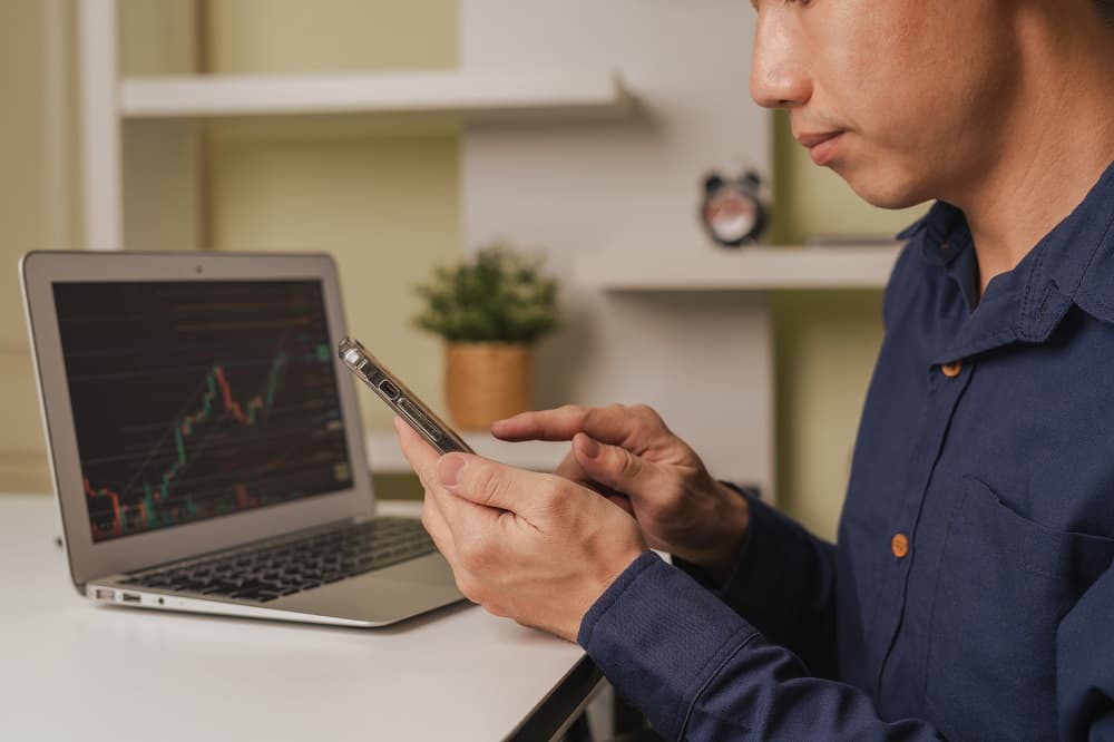 Der Anleger zeigt auf das Smartphone, verwendet die Bitcoin-Sturm-Handelsplattform, einen Laptop im Hintergrund mit einem Börsendiagramm auf dem Bildschirm.